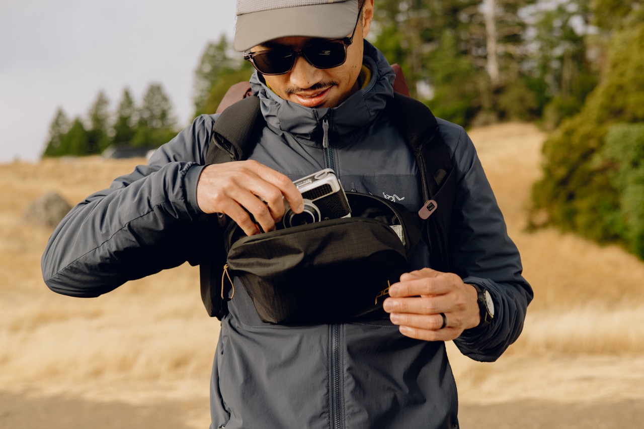 Peak Design Outdoor Sling Umhängetasche in der Farbe Schwarz, Lifestyle Foto mit Tasche als Fronttasche auf Person die gerade etwas herausnimmt mit einem Wald im Hintergrund