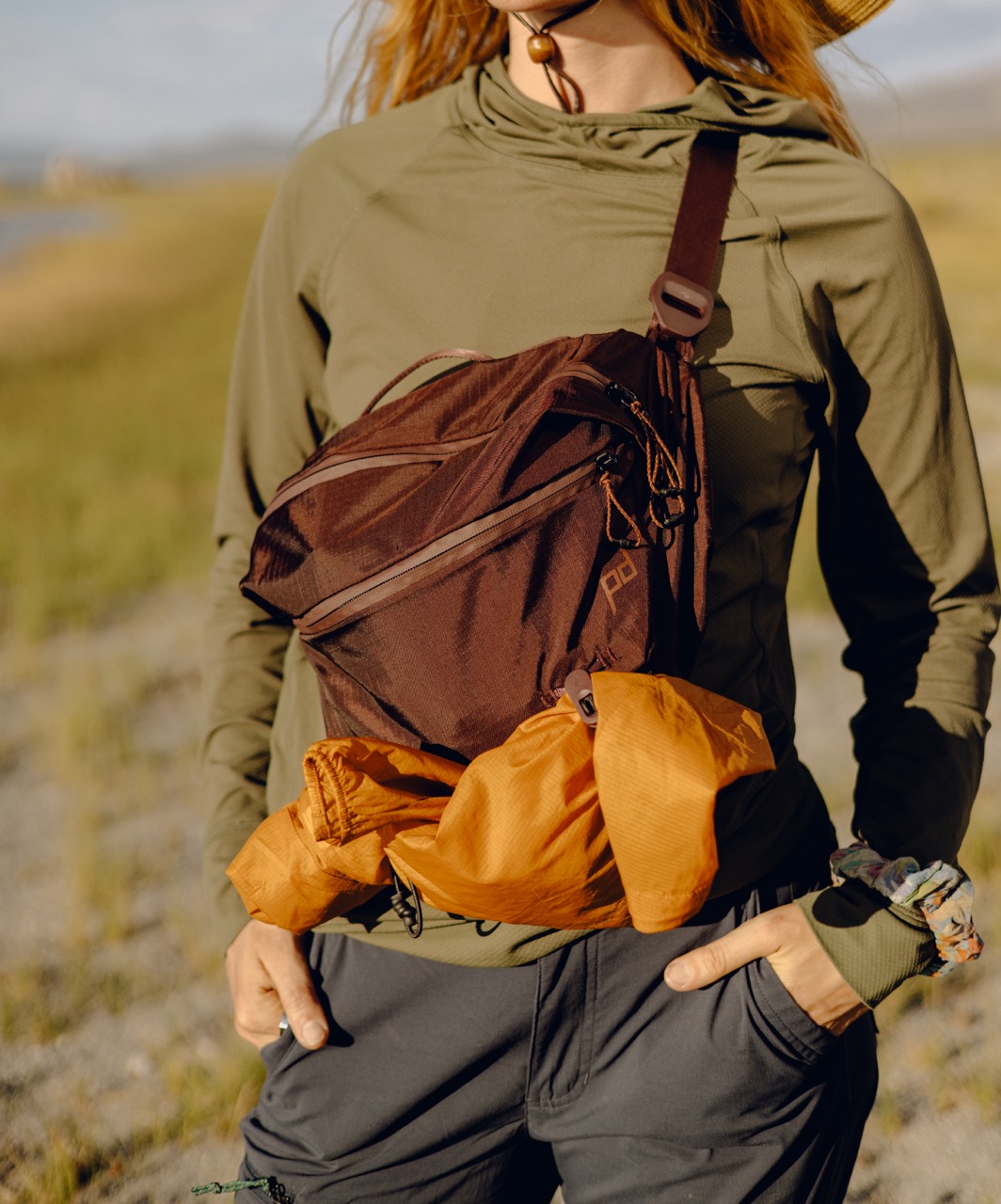 Peak Design Outdoor Sling Umhängetasche mit 7L Volumen in der Farbe Lila, Lifestyle Foto mit Tasche als Frontale Umhängetasche auf Person mit angehängter Regenjacke 