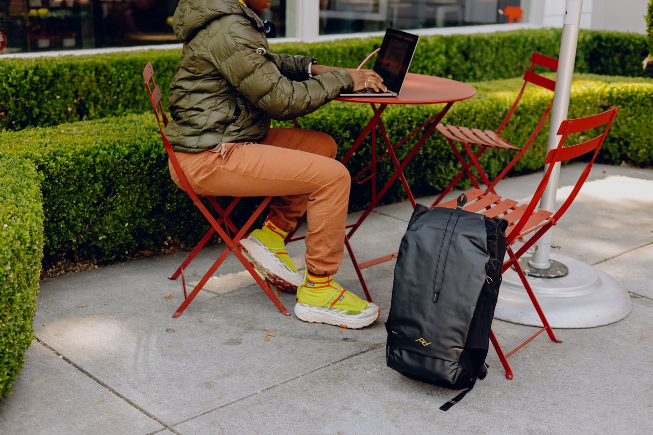 Peak Design Outdoor Backpack mit 45 Litern Fassungsvermögen in der Farbe Schwarz,  Lifestyle Foto mit Rucksack angelehnt an Sessel an einem Tisch an dem eine Person sitzt