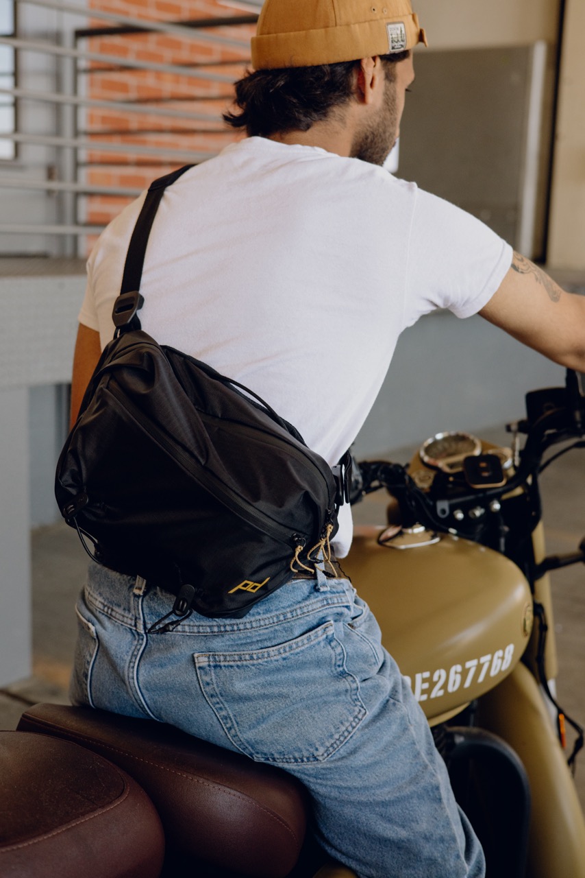 Peak Design Outdoor Sling Umhängetasche mit 7L Volumen in der Farbe Schwarz, Lifestyle Foto mit Tasche als Umhängetasche auf Rücken einer Person die auf einem Motorrad sitzt